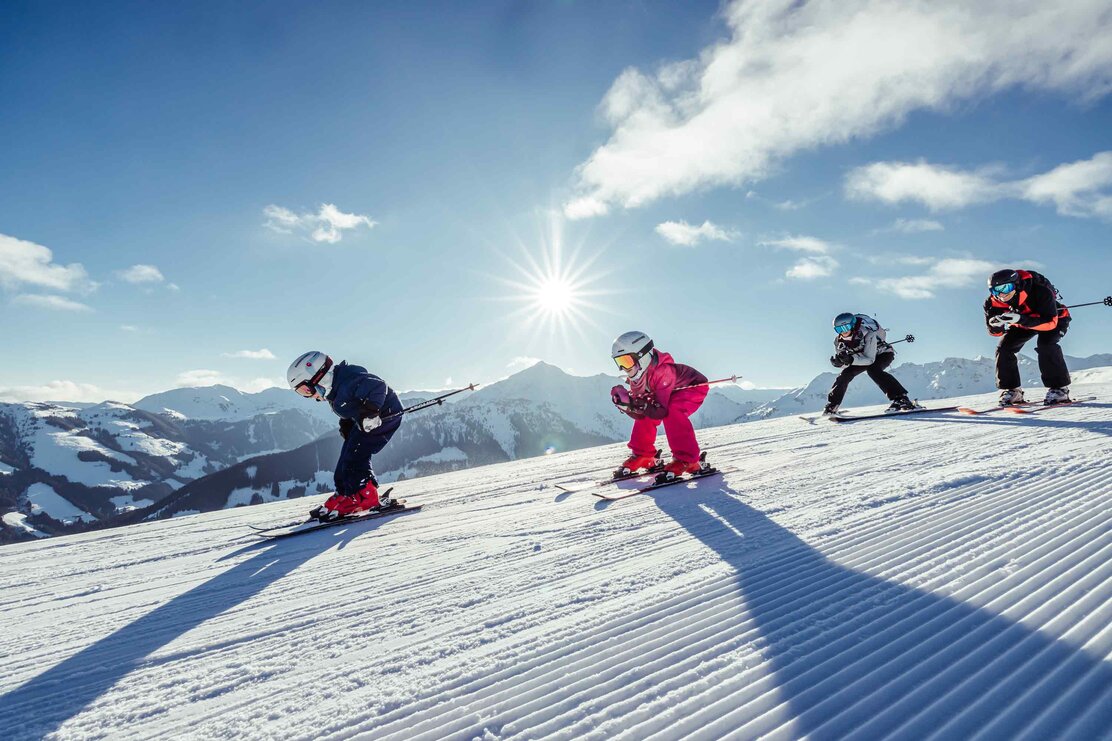 Eine Familie fährt gemeinsam in Hocke die Skipiste runter, im Hintergrund zu sehen die winterliche Berglandschaft | © Alpbachtal Tourismus | shootandstyle 