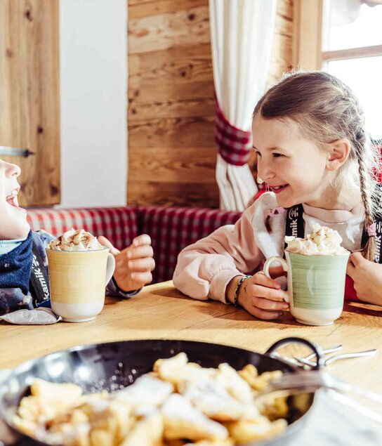 Zwei Kinder sitzen in der Skihütte, trinken heiße Schokolade mit Sahne und haben vor sich eine Pfanne voller Kaiserschmarrn stehen | © Alpbachtal Tourismus | shootandstyle 