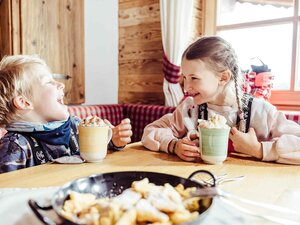 Zwei Kinder sitzen in der Skihütte, trinken heiße Schokolade mit Sahne und haben vor sich eine Pfanne voller Kaiserschmarrn stehen | © Alpbachtal Tourismus | shootandstyle 