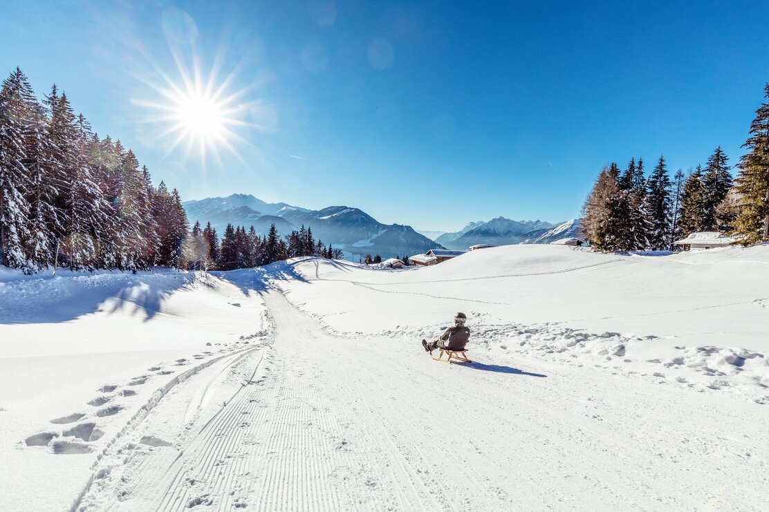 Ein Mann rodelt an der Rodelbahn am Reither Kogel entlang, um ihn eine verschneite Winterlandschaft, über ihm die strahlende Sonne 