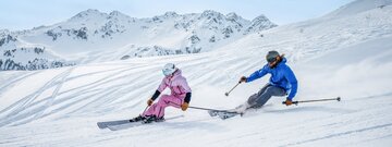 Ein Mann und eine Frau auf der Piste beim Skifahren mit Panorama im Hintergrund. | © Ski Juwel Alpbachtal Wildschönau | Christoph Oberschneider