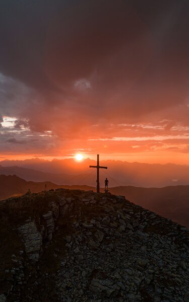Man sieht ein Gipfelkreuz auf einem Berg mit einem Menschen der den Sonnenaufgang beobachtet. | © Alpbachtal Tourismus