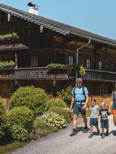 Eine Familie wandert der Straße entlang, vorbei an einem schönen alten Bauernhaus, welches umgeben ist von blühender Natur  | © Alpbachtal Tourismus | Mathäus Gartner 