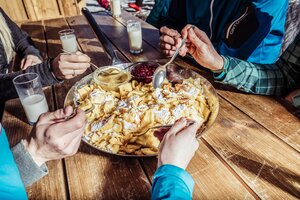 Nahaufnahme einer Portion Kaiserschmarren, angerichtet in einer großen Pfanne mit Apfelmus und Preiselbeeren, nebenan ein paar Gläser Milch | © Ski Juwel Alpbachtal Wildschönau | shootandstyle 