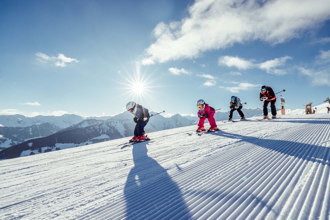 Eine Familie fährt gemeinsam in Hocke die Skipiste runter, im Hintergrund zu sehen die winterliche Berglandschaft | © Alpbachtal Tourismus | shootandstyle 