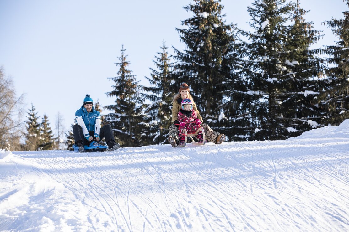 Eine Familie rodelt gemeinsam die Rodelstrecke entlang, Mutter und Tochter auf einer Rodel, der Vater nebenbei  | © Alpbachtal Tourismus | shootandstyle 