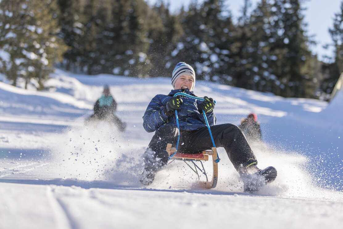 Auf diesem Bild zu sehen ist ein Junge, welcher gerade die Rodelbahn hinunter rodelt, im Hintergrund seine Familie  | © Alpbachtal Tourismus | shootandstyle 