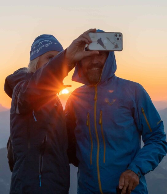 Auf diesem Bild sieht man eine Frau und einen Mann, welche ein Selfie machen, im Hintergrund der schöne Sonnenaufgang  | © Alpbachtal Tourismus | shootandstyle 